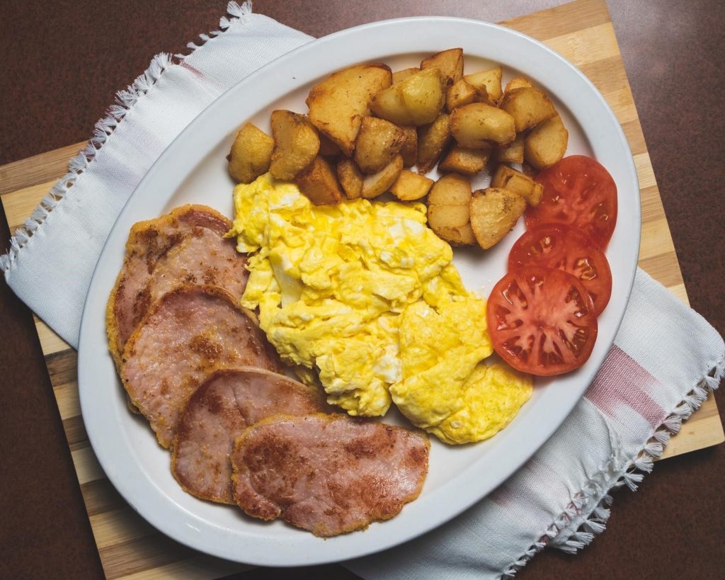Overhead shot of a huge plate with peameal bacon and scrambled eggs with home fries and sliced tomatoes.