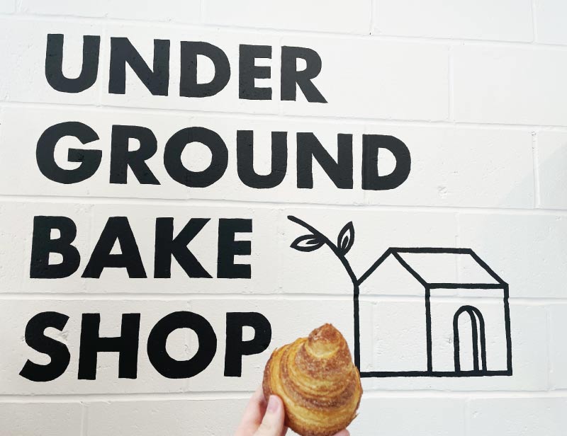 Holding a baked good in front of the Underground Bake Shop logo painted on a a wall.