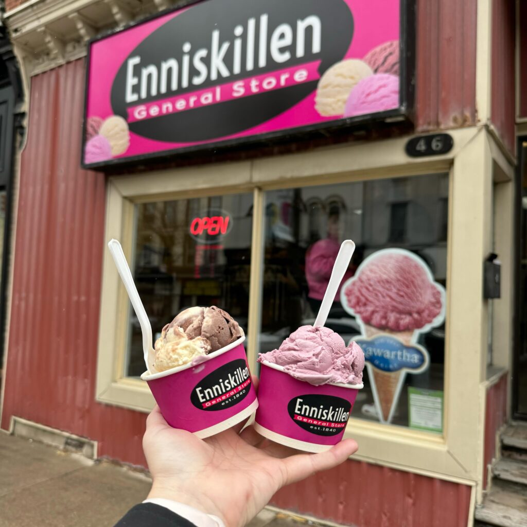 Close up of someone's hand holding two cups of ice cream with spoons sticking out in front of Enniskillen General Store in downtown Bowmanville.
