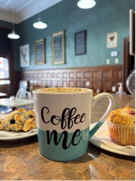 Close up of a mug that says, "Coffee me", with baked goods in the background.