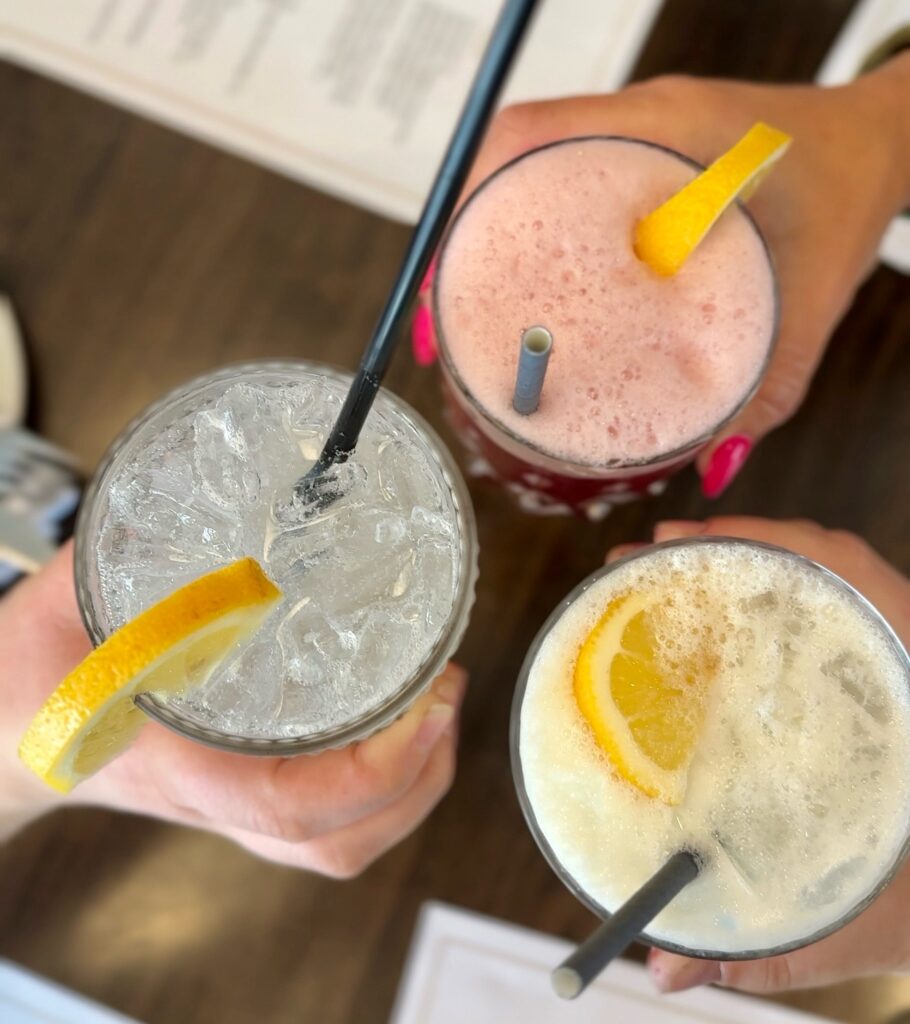 Overhead shot of three different coloured mocktails on a table with straws and lemon wedges.