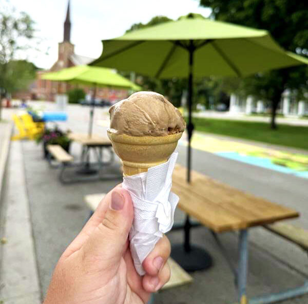 Holding a chocolate ice cream cone outside with picnic tables and umbrellas in the background.