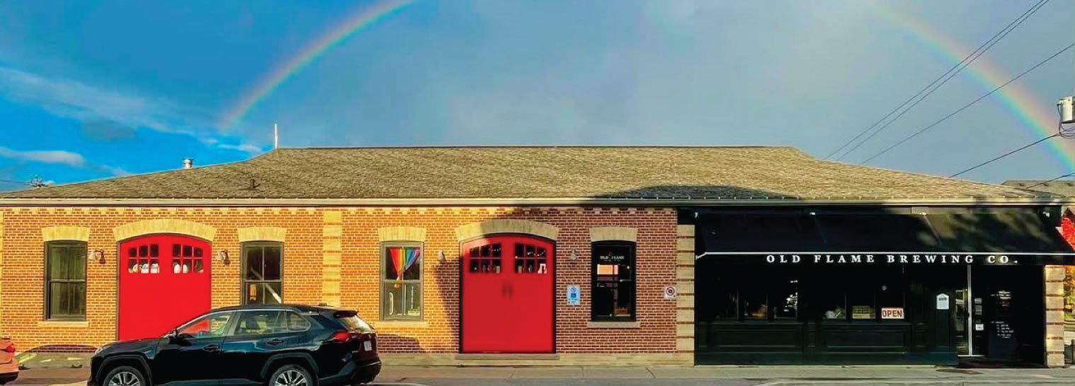 Wide shot of the historic Old Flame Brewery building on a sunny day with a rainbow in a blue sky in the background.